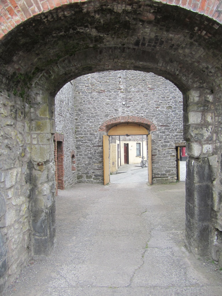Image of the exit from Elizabeth Fort, Cork. A stone archway is shown in the foreground leading out to a courtyard and an arched doorway. The doors are open leading out of the fort. 